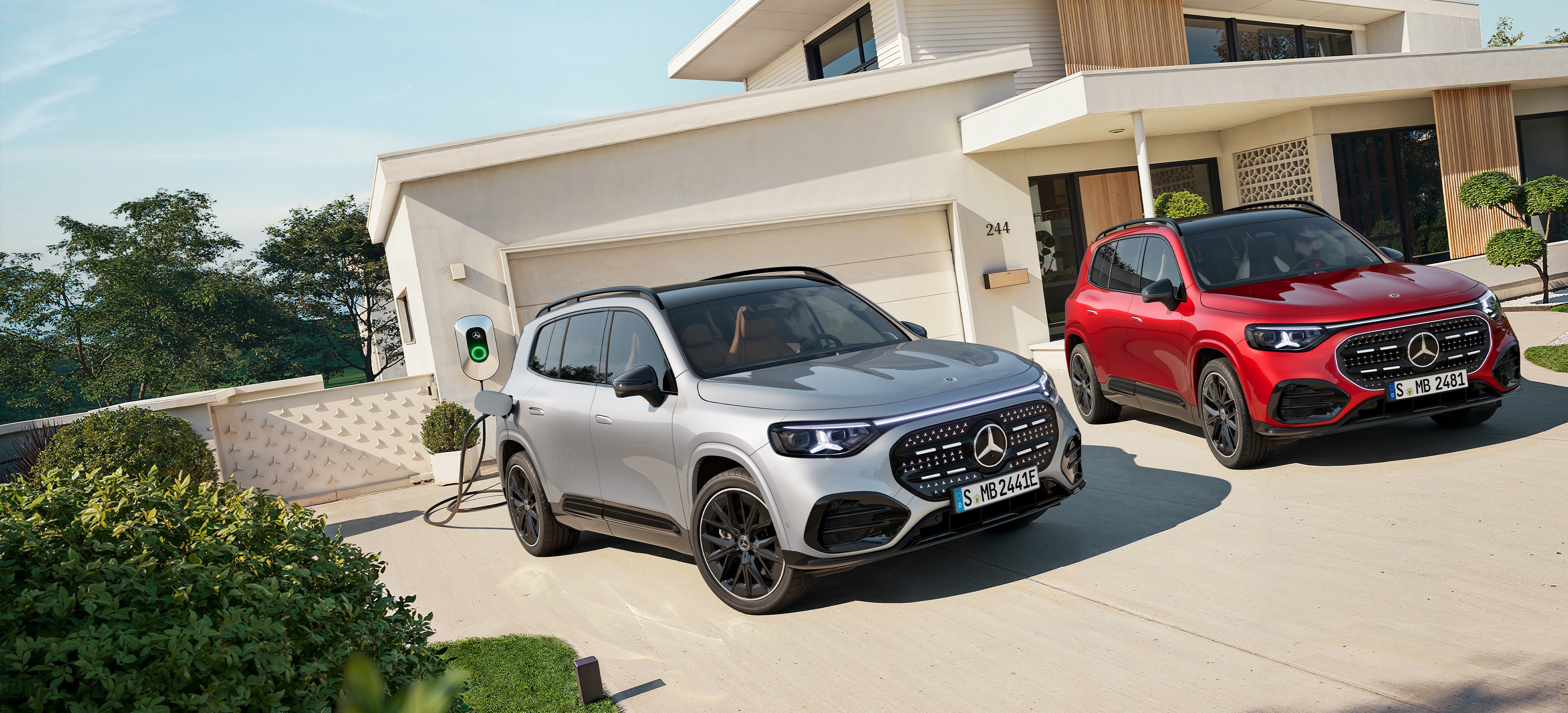 Nuevo GLB | Mercedes-Benz Un GLB eléctrico gris enchufado al cargador y otro rojo aparcados fuera de una casa