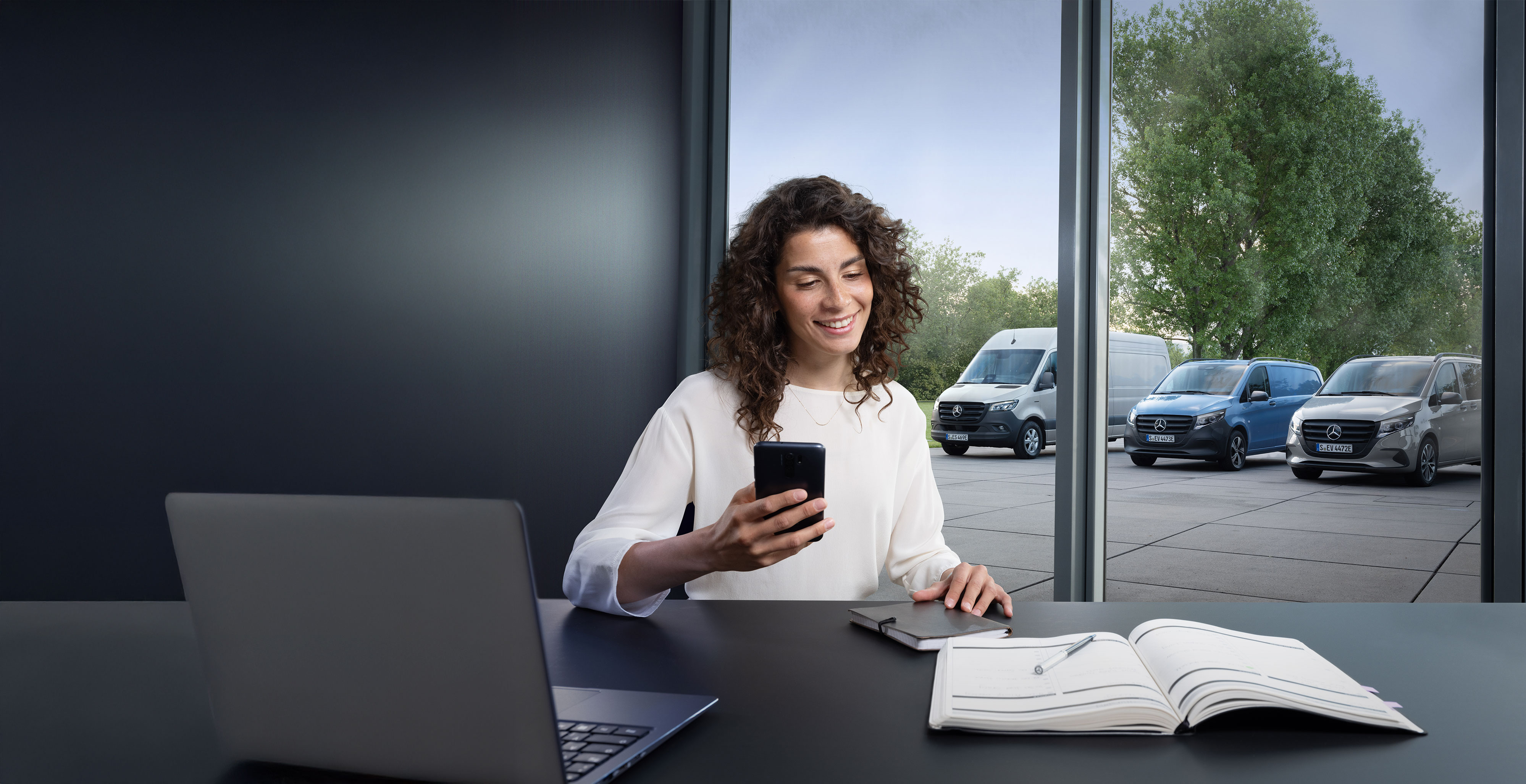 Mercedes-Benz | Conveniencia | Reserva de cita online Mujer reservando un servicio a través de smartphone con una serie de furgones Mercedes-Benz de fondo.