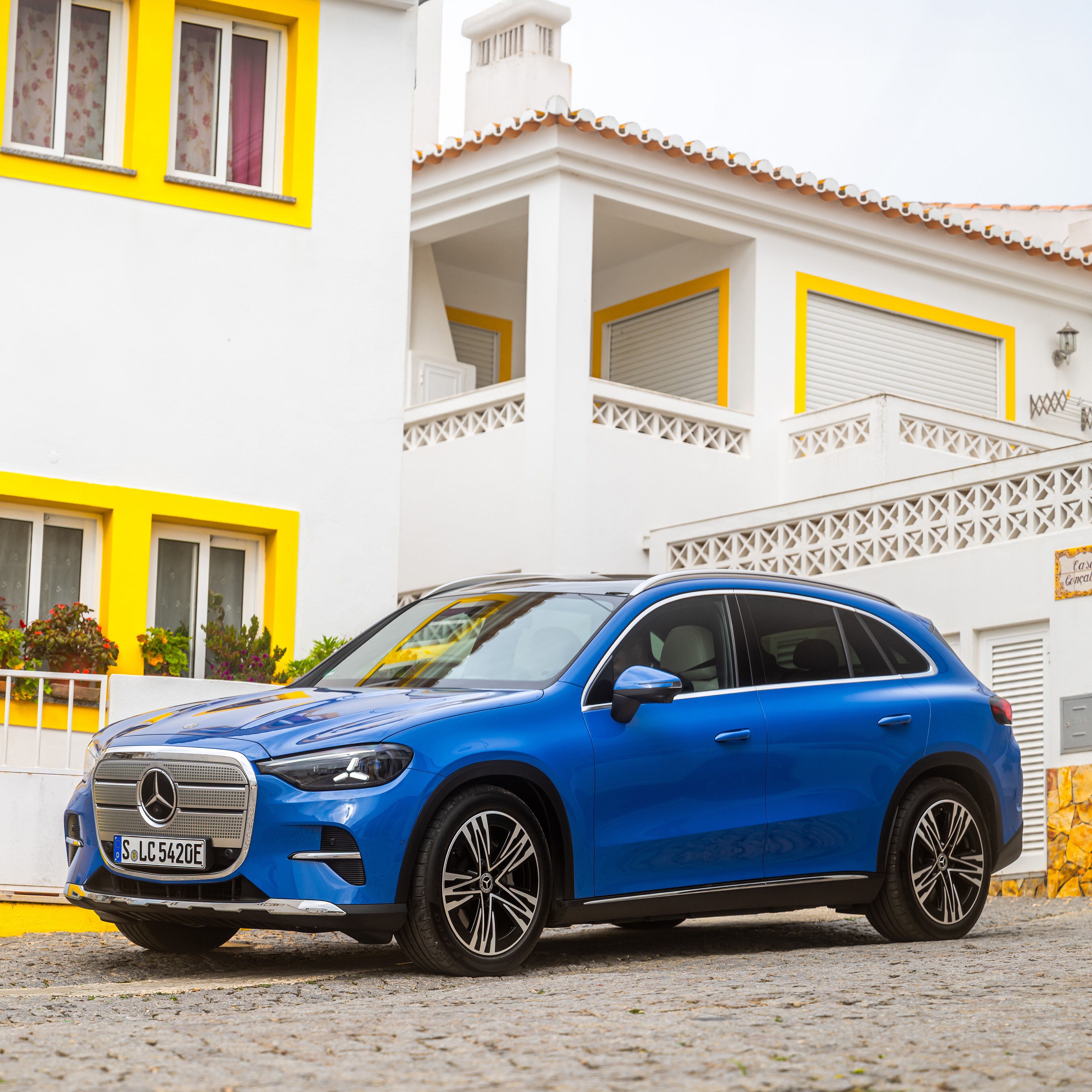 Suscríbete | Mercedes- Benz Magazine visual de un GLC en color azul en una plaza de suelo de piedra y enfrente de una casa con ventanas amarillas
